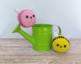 May include: A green metal watering can with two crocheted bee toys. One bee is pink with white wings, and the other is yellow with black stripes and white wings. The toys have black eyes and are on a wooden surface.