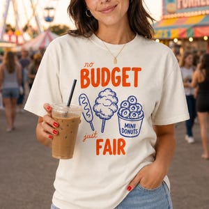 May include: A cream-colored short-sleeve t-shirt featuring the orange text "no BUDGET just FAIR" and blue line-art illustrations of a corn dog, cotton candy, and a bucket labeled "MINI DONUTS" centered on the front of the garment.