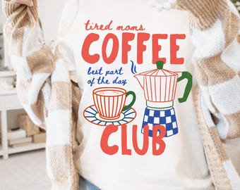 May include: A cream-colored crewneck sweatshirt featuring a graphic print with the red text "tired moms COFFEE CLUB" and "best part of the day" in blue. The design includes a red and white striped coffee cup and a moka pot with a blue checkered base.