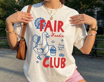 May include: A white short-sleeve t-shirt featuring a blue line-art graphic of a duck holding a bucket and cotton candy. The text reads FAIR Foodie CLUB and Every Single Year. The design includes a Ferris wheel, stars, and small circular shapes.