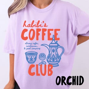May include: A light purple t-shirt featuring a graphic print in orange and blue. The text reads "habibi's COFFEE CLUB" with "strong coffee, cardamom, & good company" written in blue. The graphic includes a traditional coffee pot and cup.