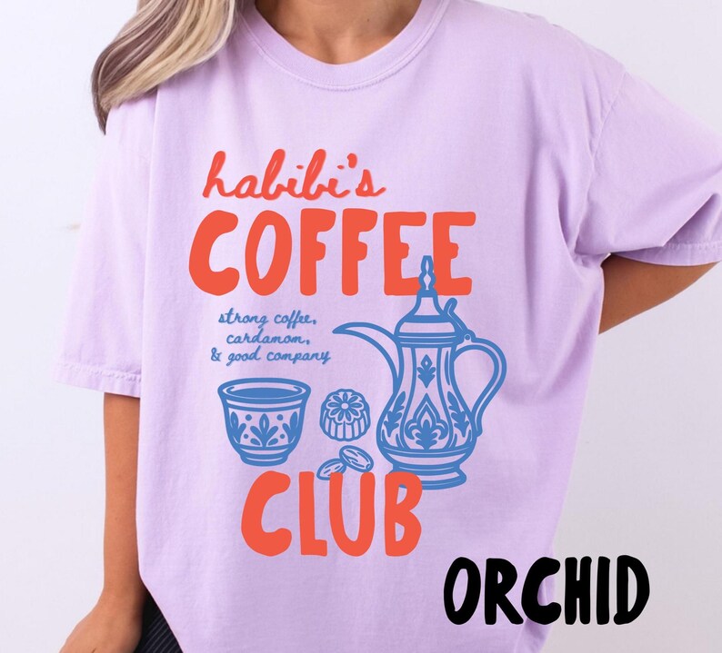 May include: A light purple t-shirt featuring a graphic print in orange and blue. The text reads "habibi's COFFEE CLUB" with "strong coffee, cardamom, & good company" written in blue. The graphic includes a traditional coffee pot and cup.