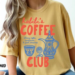 May include: A mustard yellow t-shirt featuring a graphic print in red and blue. The text reads "habibi's COFFEE CLUB" with "strong coffee, cardamom, & good company" written in blue. The design includes a blue coffee pot and a small cup.