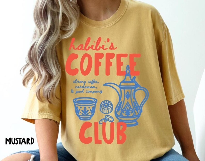 May include: A mustard yellow t-shirt featuring a graphic print in red and blue. The text reads "habibi's COFFEE CLUB" with "strong coffee, cardamom, & good company" written in blue. The design includes a blue coffee pot and a small cup.