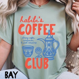 May include: A sage green t-shirt featuring the orange text "habibi's COFFEE CLUB" and a blue graphic of a traditional coffee pot, a small cup, and dates. The text "strong coffee, cardamom, & good company" is printed in blue script between the graphics.