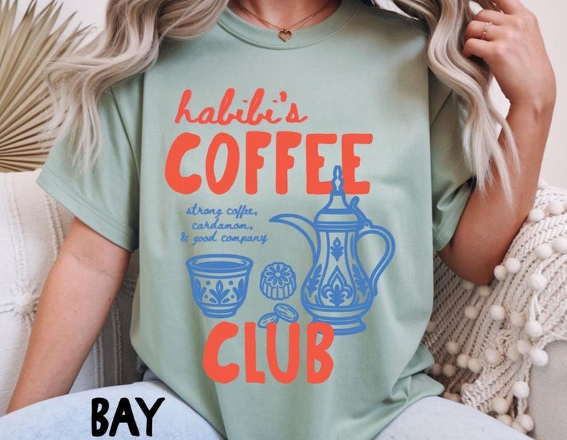 May include: A sage green t-shirt featuring the orange text "habibi's COFFEE CLUB" and a blue graphic of a traditional coffee pot, a small cup, and dates. The text "strong coffee, cardamom, & good company" is printed in blue script between the graphics.