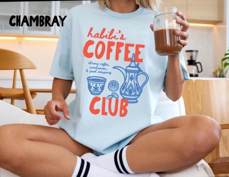 May include: A light blue t-shirt featuring the text "CHAMBRAY" in the top left corner and "habibi's COFFEE CLUB" in large red letters. The graphic includes a blue illustration of a coffee pot, a cup, and the text "strong coffee, cardamom, & good company."