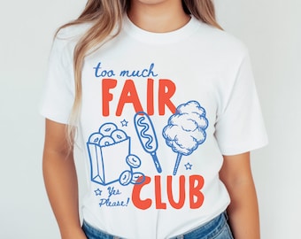 May include: A white short-sleeve t-shirt featuring a graphic print with the text "too much FAIR CLUB" and "Yes Please!". The design includes blue line art of a bag of donuts, a corn dog, and cotton candy, with red block lettering.