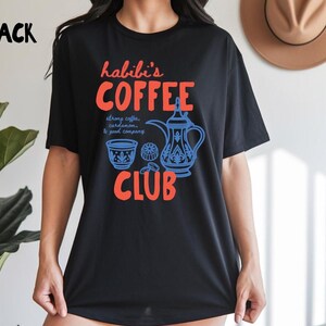 May include: A black short-sleeve t-shirt featuring a graphic print with the text "habibi's COFFEE CLUB" in orange. The design includes a blue illustration of a traditional coffee pot, a small cup, and the text "strong coffee, cardamom, & good company."