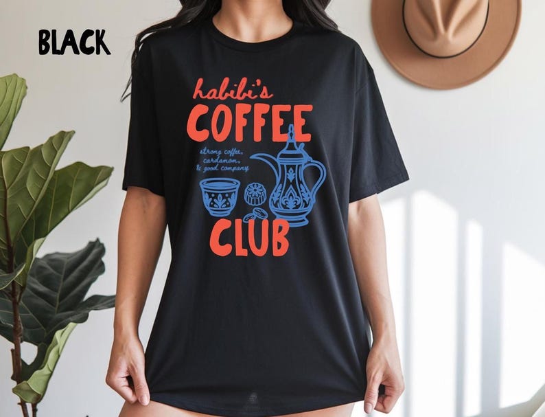 May include: A black short-sleeve t-shirt featuring a graphic print with the text "habibi's COFFEE CLUB" in orange. The design includes a blue illustration of a traditional coffee pot, a small cup, and the text "strong coffee, cardamom, & good company."