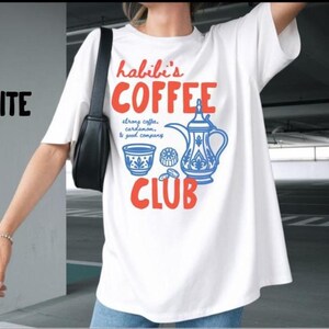 May include: A white short-sleeved t-shirt featuring a graphic print with the text "habibi's COFFEE CLUB" in orange, alongside a blue illustration of a traditional coffee pot, a small cup, and the text "strong coffee, cardamom, & good company."