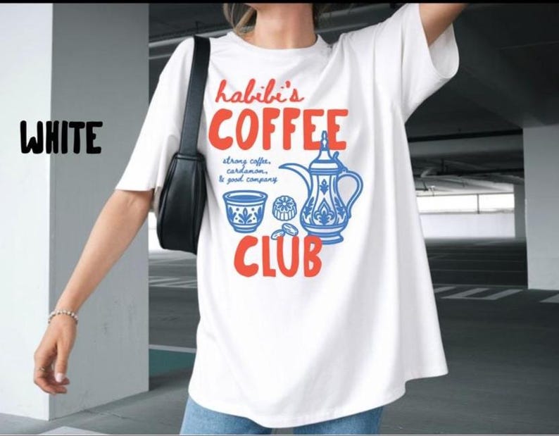 May include: A white short-sleeved t-shirt featuring a graphic print with the text "habibi's COFFEE CLUB" in orange, alongside a blue illustration of a traditional coffee pot, a small cup, and the text "strong coffee, cardamom, & good company."