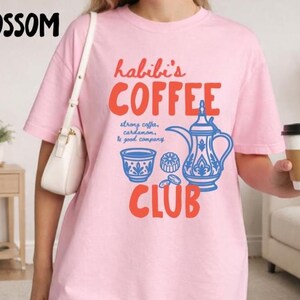 May include: A light pink t-shirt featuring the red text "habibi's COFFEE CLUB" and a blue graphic of a traditional coffee pot, a small cup, and a pastry. The text "strong coffee, cardamom, & good company" is printed in blue between the words.
