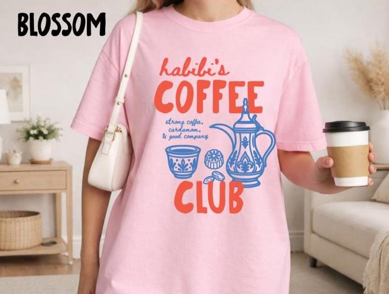 May include: A light pink t-shirt featuring the red text "habibi's COFFEE CLUB" and a blue graphic of a traditional coffee pot, a small cup, and a pastry. The text "strong coffee, cardamom, & good company" is printed in blue between the words.
