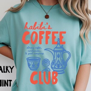 May include: A chalky mint t-shirt featuring the red text "habibi's COFFEE CLUB" and blue illustrations of a traditional coffee pot, a small cup, and cardamom pods, with the blue text "strong coffee, cardamom, & good company" printed in the center.