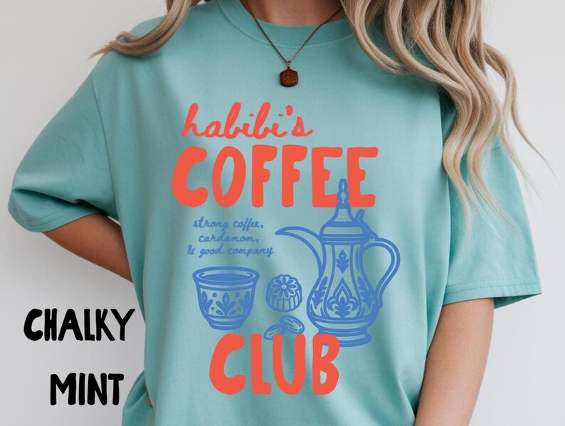 May include: A chalky mint t-shirt featuring the red text "habibi's COFFEE CLUB" and blue illustrations of a traditional coffee pot, a small cup, and cardamom pods, with the blue text "strong coffee, cardamom, & good company" printed in the center.