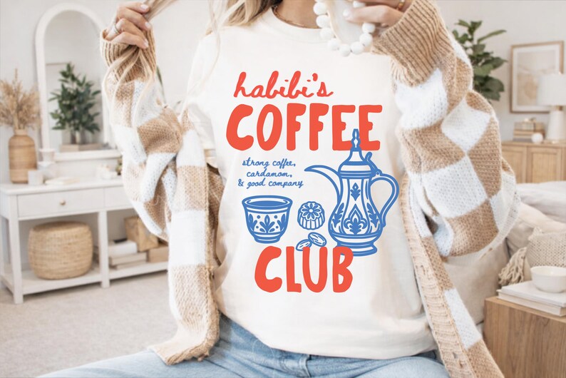 May include: A cream-colored t-shirt featuring red text that reads habibi's COFFEE CLUB and blue graphic illustrations of a traditional coffee pot, a small cup, and coffee beans with the text strong coffee, cardamom, & good company.