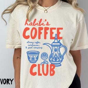 May include: An ivory short-sleeve t-shirt featuring red text that reads Habibi's COFFEE CLUB. Below the text, blue line art depicts a traditional coffee pot, a small cup, and a pastry, with the blue text strong coffee, cardamom, & good company.