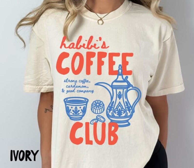 May include: An ivory short-sleeve t-shirt featuring red text that reads Habibi's COFFEE CLUB. Below the text, blue line art depicts a traditional coffee pot, a small cup, and a pastry, with the blue text strong coffee, cardamom, & good company.