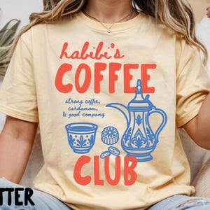 May include: A butter yellow t-shirt featuring red text that reads habibi's COFFEE CLUB and blue graphic illustrations of a traditional coffee pot, a small cup, and coffee beans with the text strong coffee, cardamom, & good company.
