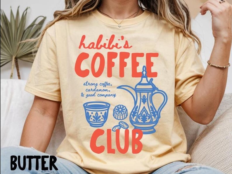 May include: A butter yellow t-shirt featuring red text that reads habibi's COFFEE CLUB and blue graphic illustrations of a traditional coffee pot, a small cup, and coffee beans with the text strong coffee, cardamom, & good company.