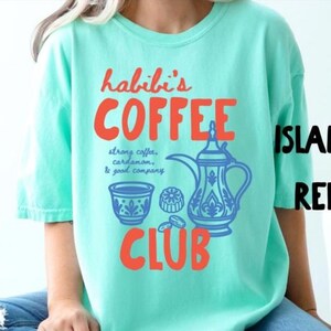 May include: A mint green t-shirt featuring a graphic print with the orange text "habibi's COFFEE CLUB" and a blue illustration of a traditional coffee pot, a small cup, and coffee beans with the text "strong coffee, cardamom, & good company."