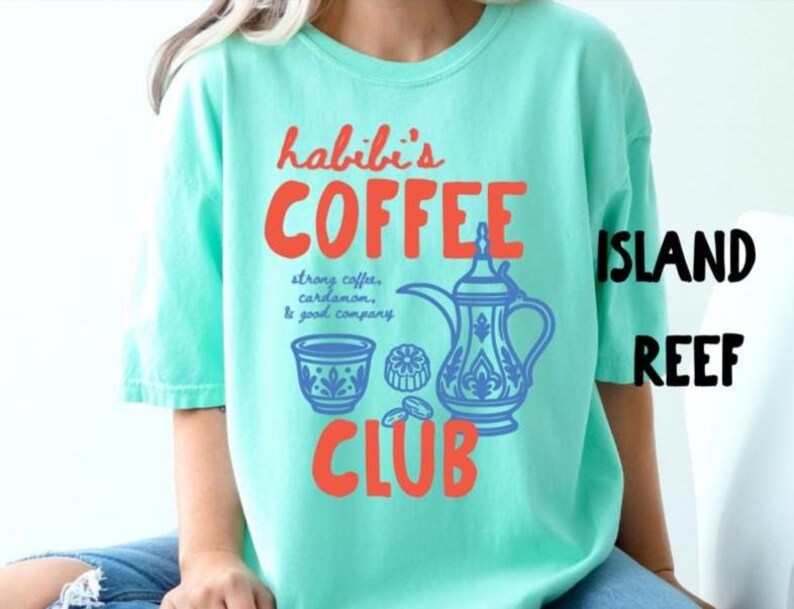 May include: A mint green t-shirt featuring a graphic print with the orange text "habibi's COFFEE CLUB" and a blue illustration of a traditional coffee pot, a small cup, and coffee beans with the text "strong coffee, cardamom, & good company."