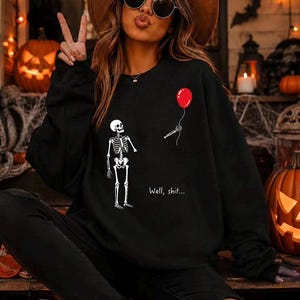 May include: A black long-sleeve sweatshirt featuring a white graphic of a skeleton looking at a red balloon held by a detached skeletal hand. The text "Well, shit..." is printed in white below the balloon. The design is centered on the chest.