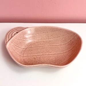 May include: A pink ceramic serving dish with a speckled texture and a handle. The dish has a curved shape with a wavy edge and horizontal lines. The handle is a rounded, stacked design.