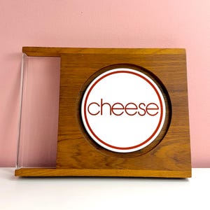 May include: A rectangular wooden serving board featuring a circular white ceramic insert in the center. The word cheese is printed in red lowercase letters on the insert, which is surrounded by a thin red circular border. A clear acrylic side panel.