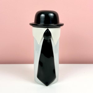 May include: A white ceramic mug with a black bowler hat and a black tie. The mug has a white collar and a white shirt design.