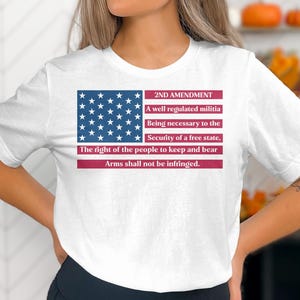 Può includere: T-shirt bianca con grafica della bandiera americana e testo del 2° emendamento. Il design include le parole "2ND AMENDMENT" e il testo "Arms shall not be infringed."