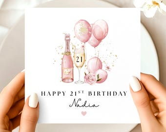 May include: A white greeting card with a watercolor illustration of a pink champagne bottle, champagne glass with the number 21, and pink balloons. The text reads "HAPPY 21ST BIRTHDAY Nadia" with a small pink heart.