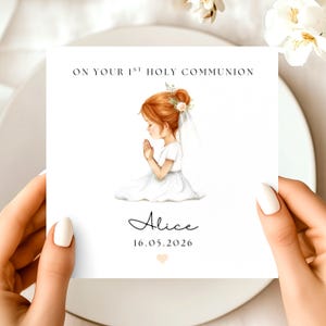 May include: A square card with the text "ON YOUR 1ST HOLY COMMUNION" and the name "Alice" with the date "16.05.2026". The card features an illustration of a girl in a white dress with red hair, praying. The card is held by two hands.