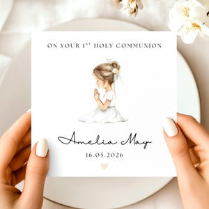May include: A white card with the text "ON YOUR 1ST HOLY COMMUNION" and the name "Amelia May" in cursive. A watercolor illustration of a girl in a white dress praying is featured. The date "16.05.2026" is also included.