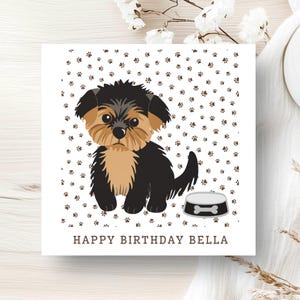 May include: A square greeting card featuring a cartoon illustration of a black and tan puppy sitting next to a grey food bowl. The background is white with a pattern of small brown paw prints. The text HAPPY BIRTHDAY BELLA is printed in brown.