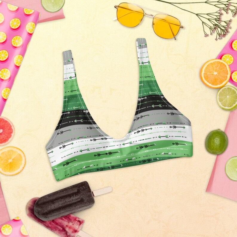 May include: A green, black, and gray striped bikini top with a pattern of arrows. The bikini top is on a light yellow background with fruit and sunglasses.