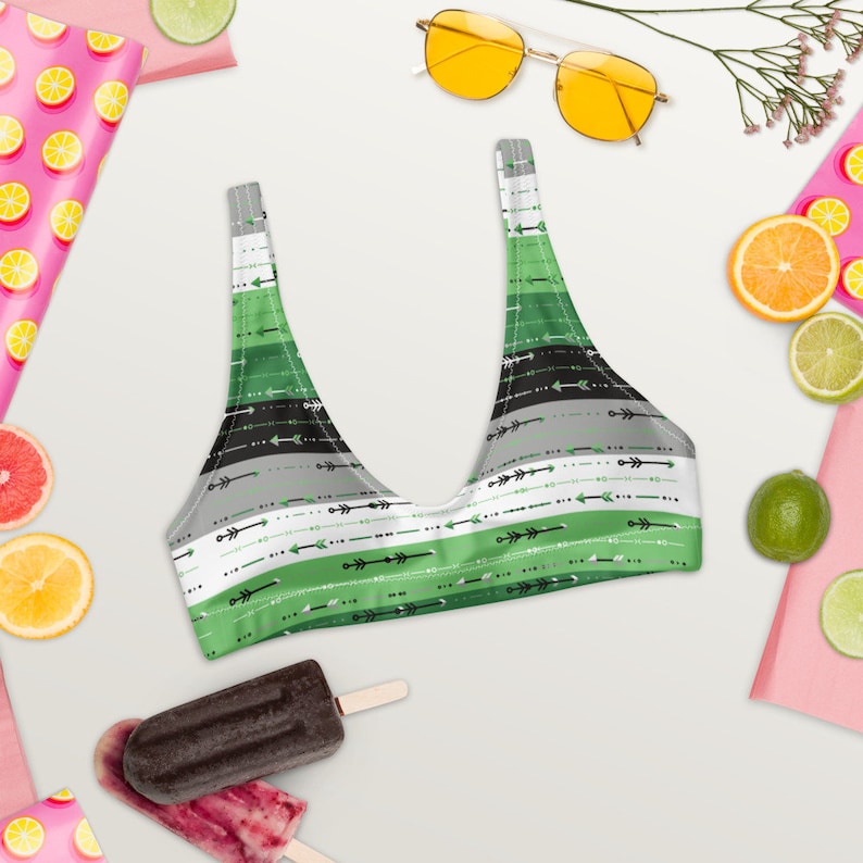 May include: Green, black, and white striped bikini top with a pattern of arrows. The bikini top is on a white background with fruit and sunglasses.