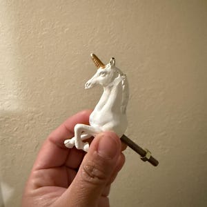 May include: White unicorn cabinet knob with a gold horn. The unicorn is in a rearing pose, attached to a threaded rod with a nut. The knob is likely made of ceramic or resin.
