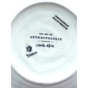 Pode incluir: Prato de cer&acirc;mica branco com o texto "THE ART OF ANTHROPOLOGIE" e "Canole akins" impresso em preto. O prato tamb&eacute;m tem o texto "Bianca" e "DISHWASHER & MICROWAVE SAFE MADE IN CHINA / FABRIQU&Eacute; EN CHINE".