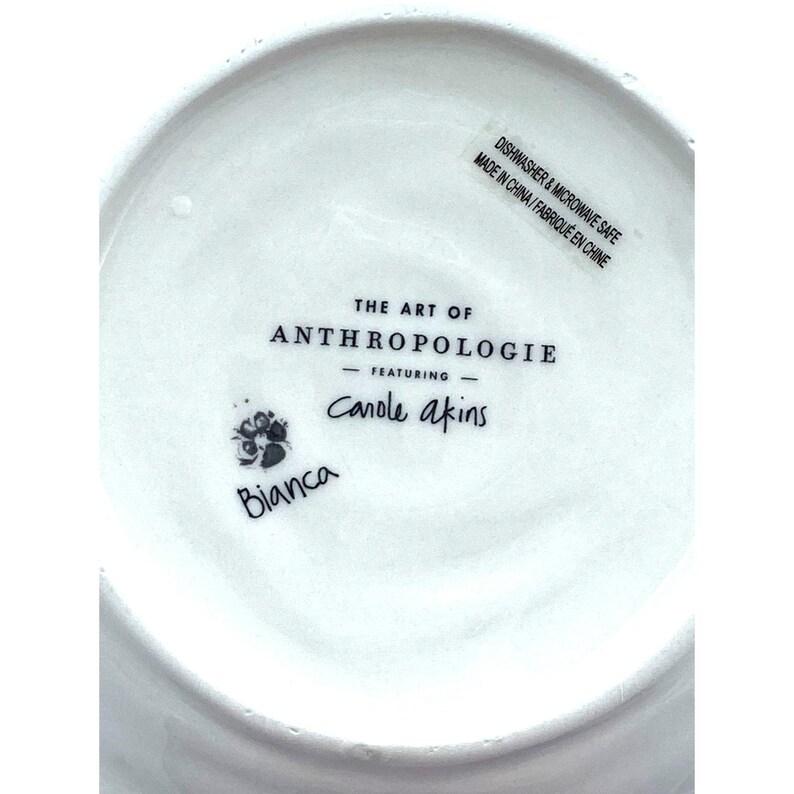 Pode incluir: Prato de cer&acirc;mica branco com o texto "THE ART OF ANTHROPOLOGIE" e "Canole akins" impresso em preto. O prato tamb&eacute;m tem o texto "Bianca" e "DISHWASHER & MICROWAVE SAFE MADE IN CHINA / FABRIQU&Eacute; EN CHINE".