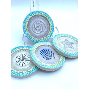 May include: Set of four round ceramic coasters with a blue and green mosaic design. Each coaster features a different sea creature in the center: a nautilus, a starfish, a seashell, and a sea urchin.