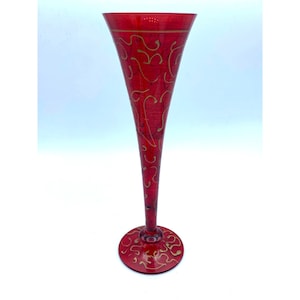 May include: A tall, red glass champagne flute with a gold swirl design. The flute has a wide base and a narrow top.