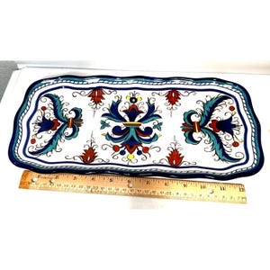 May include: A rectangular ceramic serving tray with a white background and a hand-painted floral pattern in blue, teal, and red. The tray features a scalloped edge and is positioned above a wooden ruler showing a length of approximately 12 inches.