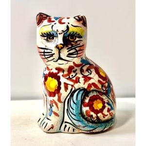 May include: A ceramic cat figurine with a colorful floral design. The cat is white with red, yellow, and blue accents. The figurine has a crackle finish and is sitting upright.