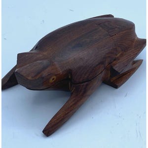 May include: A wooden frog-shaped box with a hinged lid. The box is made of dark brown wood and has a natural wood finish. The frog's eyes are made of a light-colored wood.