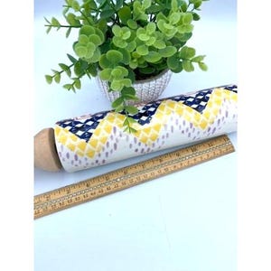 May include: A white ceramic rolling pin with a yellow, blue, and purple geometric pattern. The rolling pin has a wooden handle and is sitting on a wooden ruler.