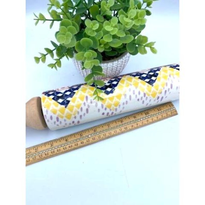 May include: A white ceramic rolling pin with a yellow, blue, and purple geometric pattern. The rolling pin has a wooden handle and is sitting on a wooden ruler.