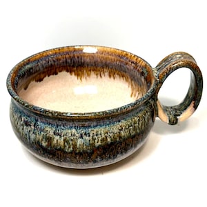 May include: A ceramic soup mug featuring a wide, rounded bowl shape and a single side handle. The exterior displays a reactive glaze with blended tones of deep blue, forest green, and earthy brown, while the interior has a smooth, cream-colored finish.