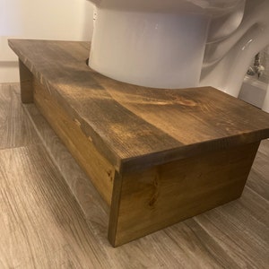 May include: A stained wooden toilet stool with a curved cutout to fit around the toilet base. The stool is rectangular with a flat top and a lower step. The wood has a warm, brown tone.